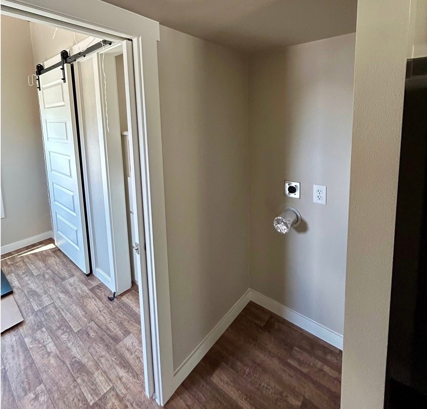Small laundry area with hookups for washer and dryer on a beige wall. Adjacent is a room with light wood flooring and a sliding white door.