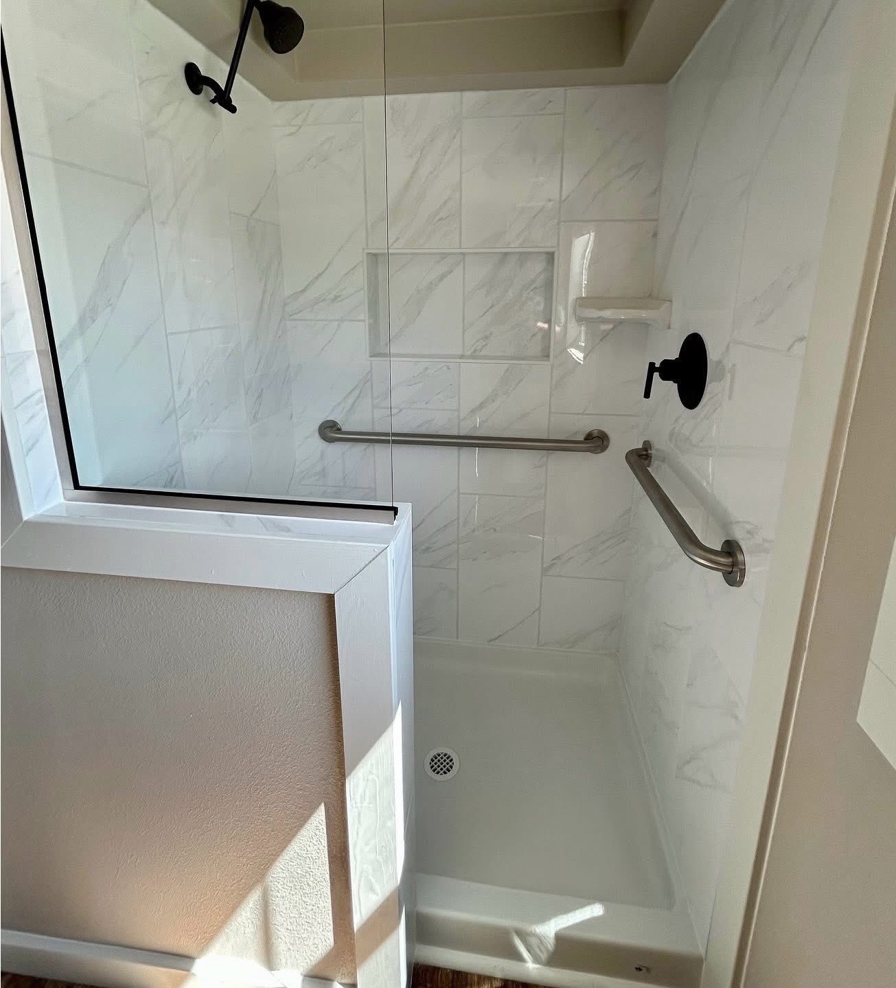 A small, white, marble-tiled shower with a glass door, black showerhead, two metal grab bars, and built-in shelf, conveying a clean, modern feel.