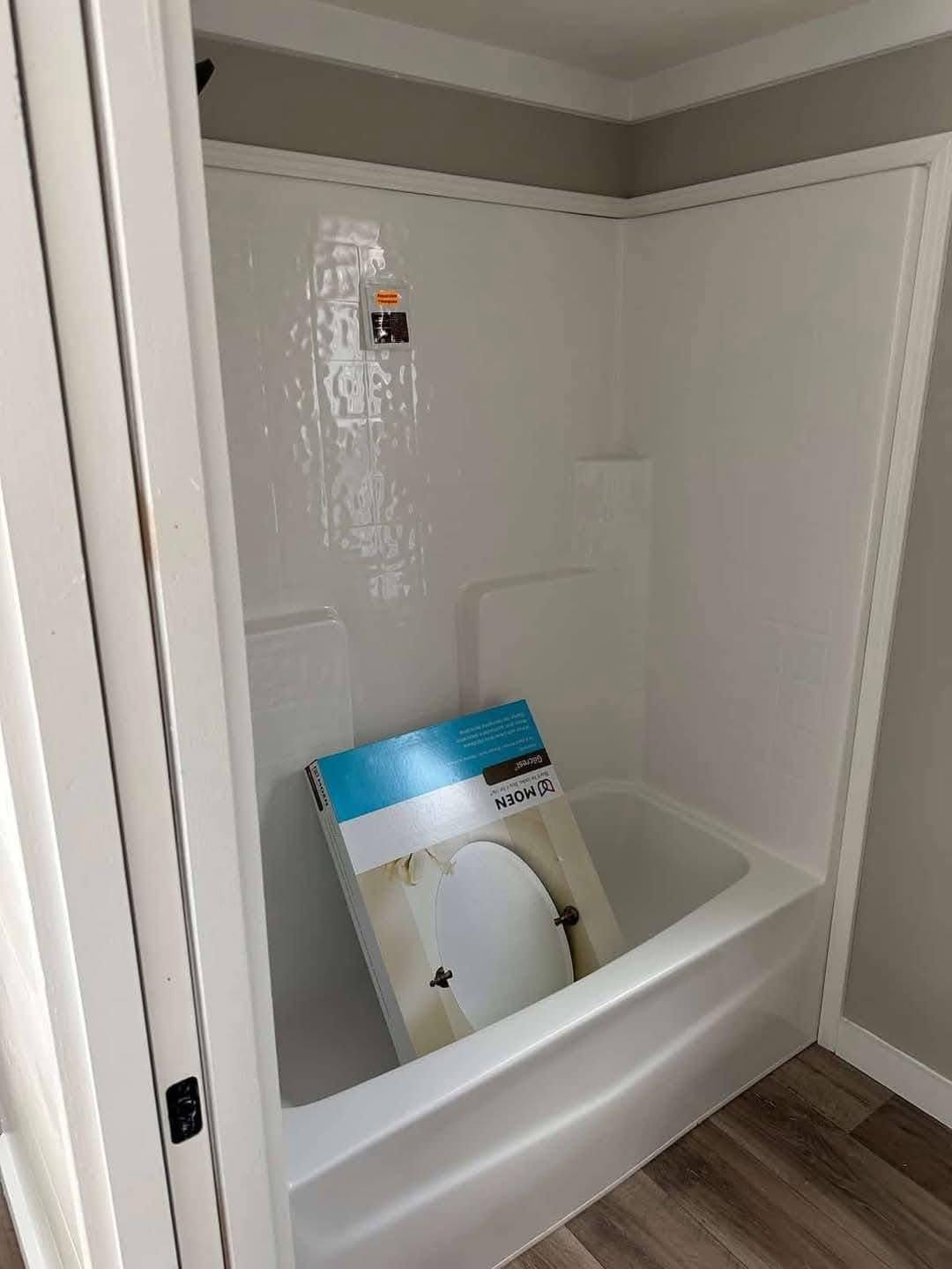A bathtub with a white surround in a small bathroom, holding an upright boxed circular mirror. Laminate wood flooring contrasts against white walls.