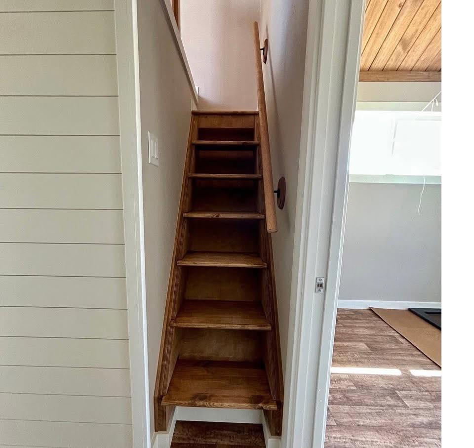 Narrow wooden staircase with steep steps leading upward between white walls. Adjacent room with wood flooring and sunlight streaming through a window.