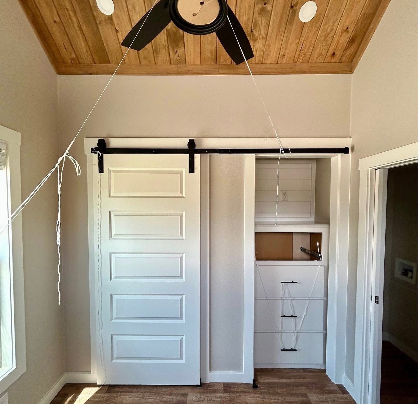 A room with a wooden ceiling and a black ceiling fan. A white sliding door on a black rail reveals a closet with shelves and cabinets.