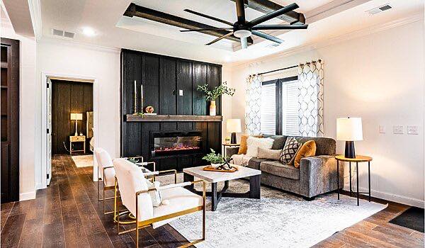 Modern living room with dark wood flooring, a gray couch, and two white chairs. A black fireplace and large ceiling fan add a cozy yet contemporary feel.