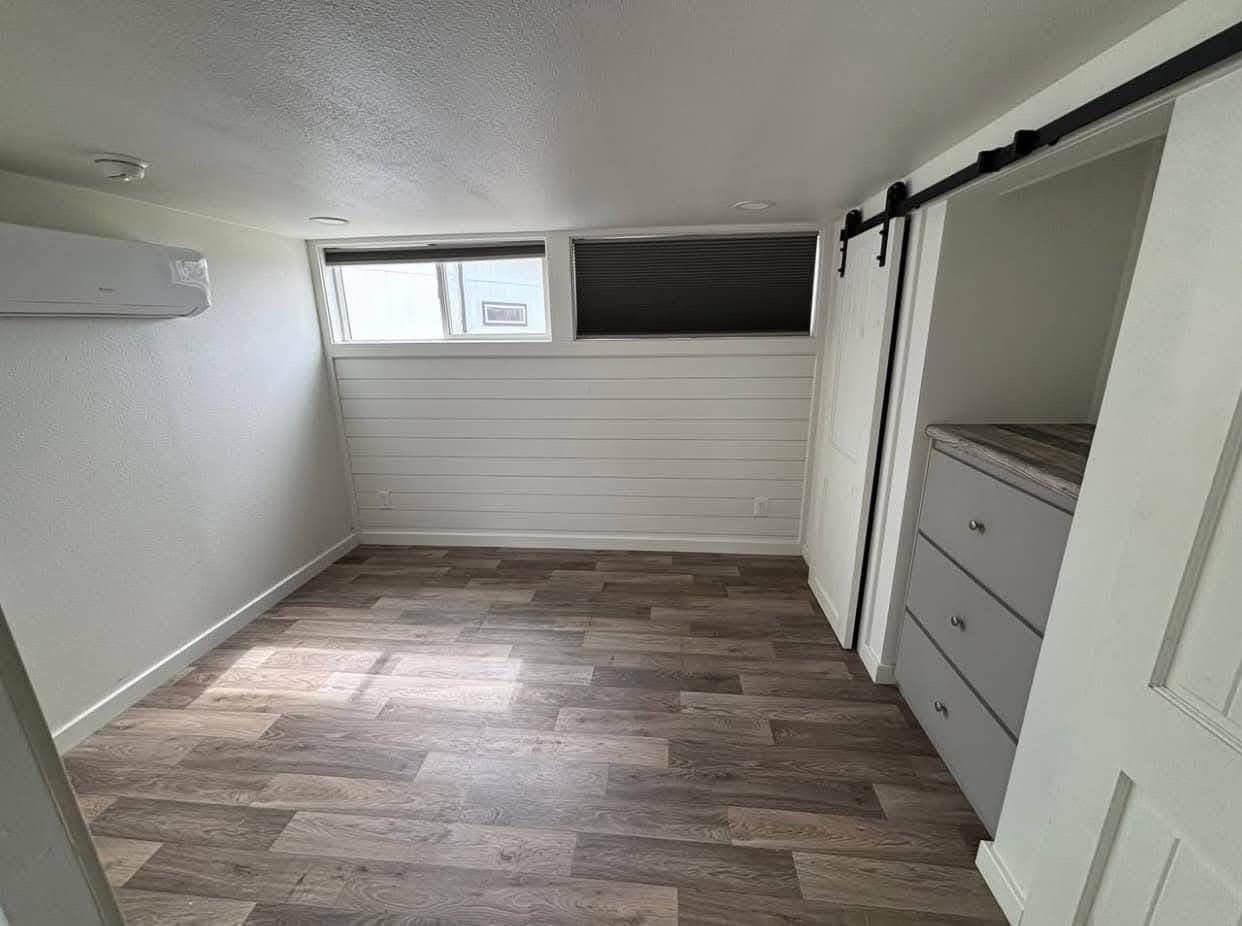 A small room with wooden flooring, white walls, and horizontal windows. It features a built-in air conditioner and a sliding barn door on the closet.