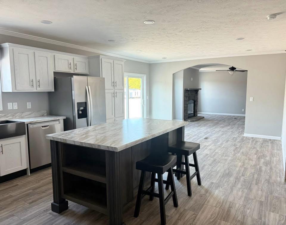Spacious kitchen with light wood floors, white cabinets, stainless steel appliances, and large island with stools. Adjacent living area with fireplace. Bright and modern.