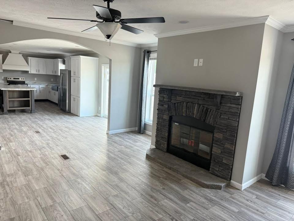 Spacious living area with gray walls, wood flooring, and a stone fireplace. A ceiling fan is overhead, leading to a modern kitchen with white cabinets.
