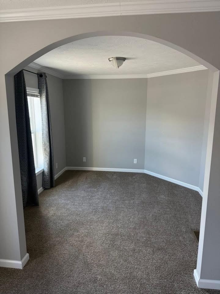 An empty, gray-carpeted room with light gray walls is visible through an archway. Natural light enters through a window with dark curtains.