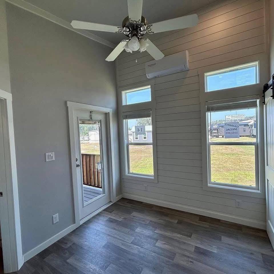 Bright, modern room with light gray walls and wood-look flooring. Features large windows, a ceiling fan, and a glass door leading outside.