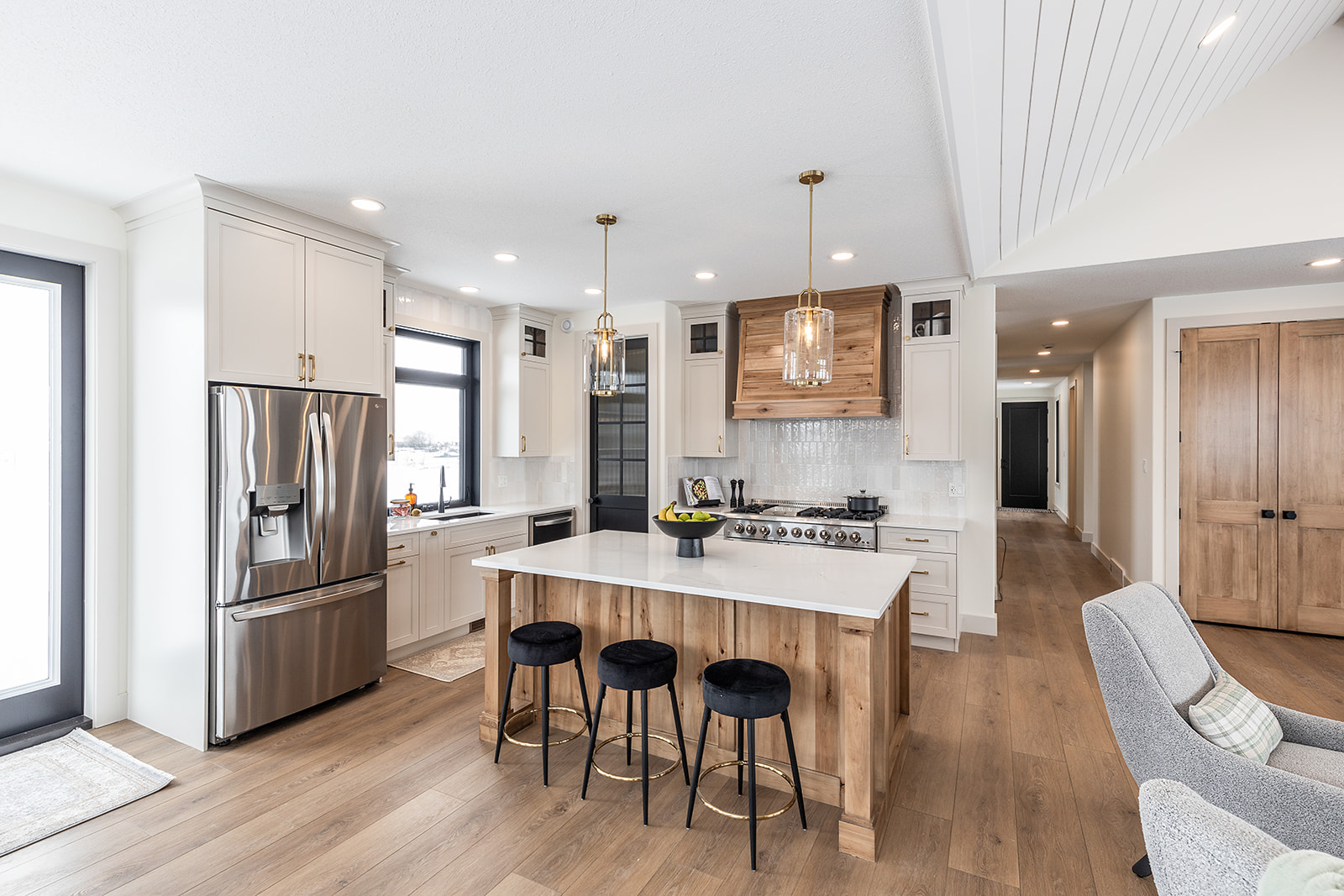 A modern kitchen with white cabinets, wooden accents, and light wood flooring. A central island with black stools and pendant lights creates a cozy ambiance.
