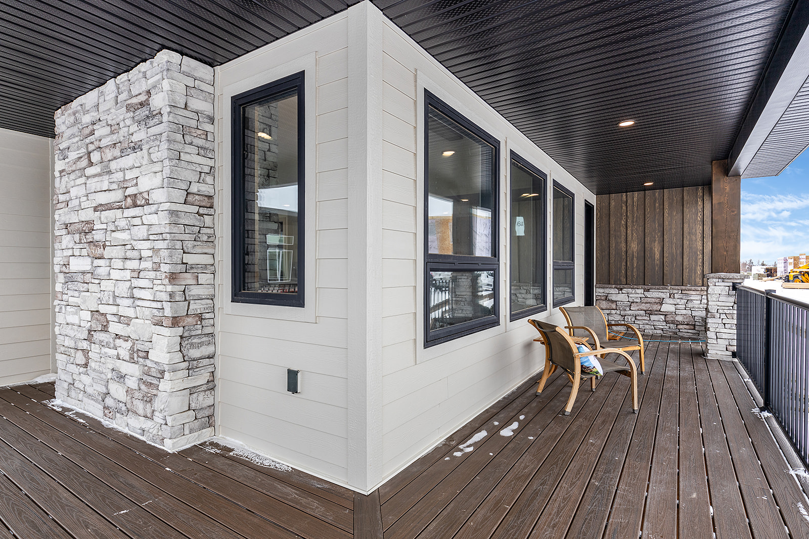 Modern outdoor deck with stone and wood siding, black-framed windows, and dark ceiling. Two chairs face a snowy landscape, creating a cozy vibe.