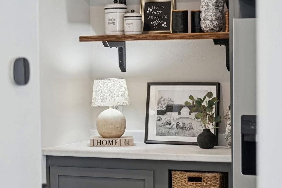 Cozy nook with a white lamp on a book labeled "Home," black-and-white photo, plants, wicker basket, and shelf with jars and a coffee sign. Warm, inviting decor.