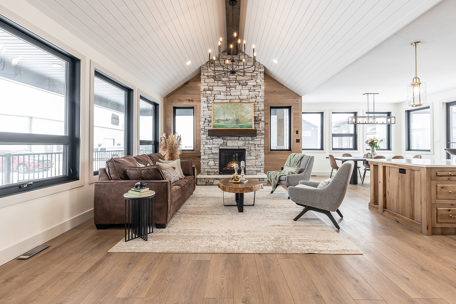 Spacious modern living room with high ceiling and chandelier. Features a stone fireplace, brown sofa, gray chairs, and wooden dining area, exuding warmth.