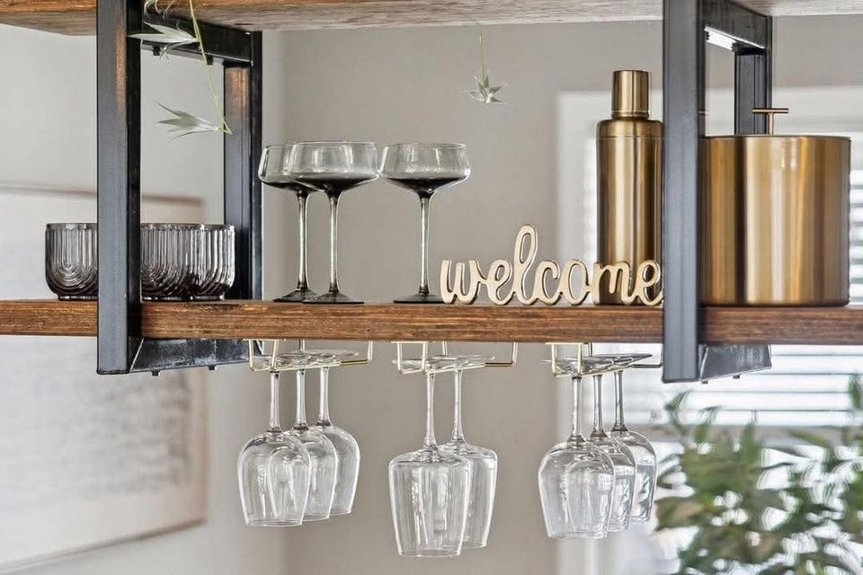 Stylish home bar setup featuring wine glasses, a gold shaker, black tumblers, and a decorative "welcome" sign on wooden shelves. Cozy and inviting atmosphere.