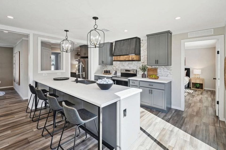 Modern kitchen with sleek gray cabinets, a large island with bar stools, and two pendant lights. Wood flooring and an open doorway to a bedroom.