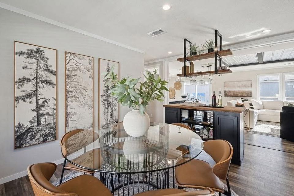 Modern dining area with a round glass table, tan leather chairs, and a white vase of greenery. Black and white tree art decorates the wall. Cozy and inviting.