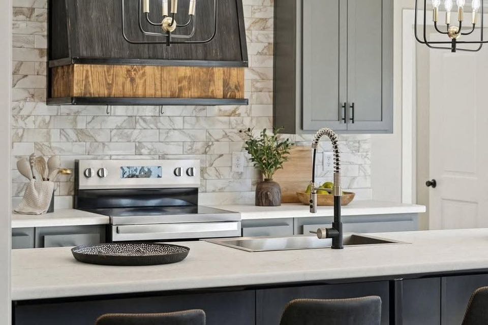 Modern kitchen with gray cabinets, marble backsplash, and an island featuring a black faucet. Elegant decor includes a plant and a decorative tray.