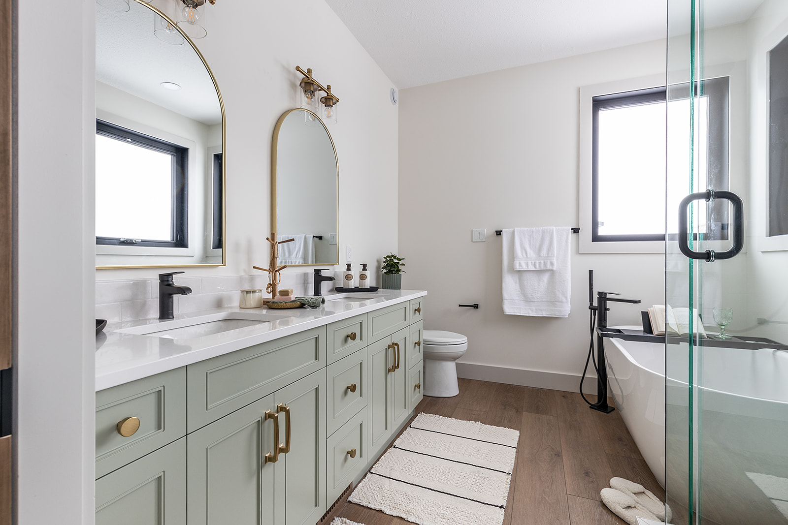 Stylish bathroom with mint green double vanity, gold accents, and arch mirrors. Features a freestanding tub, wood floor, modern decor, and bright light.