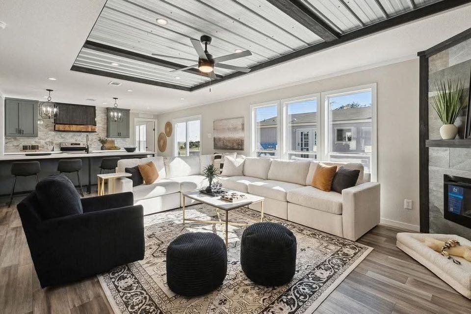 Modern living room with a white sectional sofa, black armchair, and poufs on a patterned rug. Features a sleek ceiling fan, large windows, and a cozy fireplace.