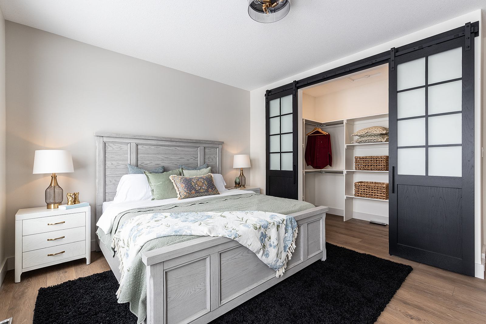 A cozy bedroom features a light gray wooden bed with floral bedding, flanked by nightstands with lamps. Black sliding doors open to a walk-in closet.