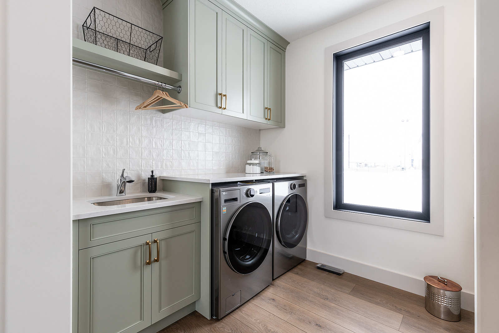 Bright laundry room with modern appliances. Green cabinets, wood floor, large window, and wall-mounted drying rod create a clean, airy feel.