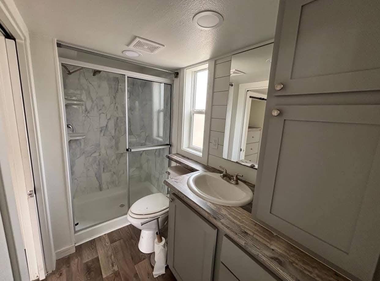 Small modern bathroom with wood floors, marble-tiled shower, sliding glass door, gray cabinets, sink, toilet, and a large mirror. Bright and clean ambiance.