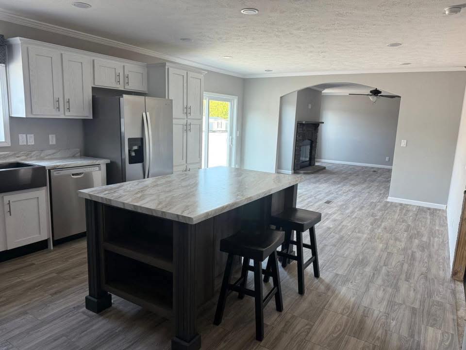 Modern kitchen with marble island, two stools, stainless steel appliances, and light wood flooring. Opens into a living room with a fireplace. Bright and inviting atmosphere.