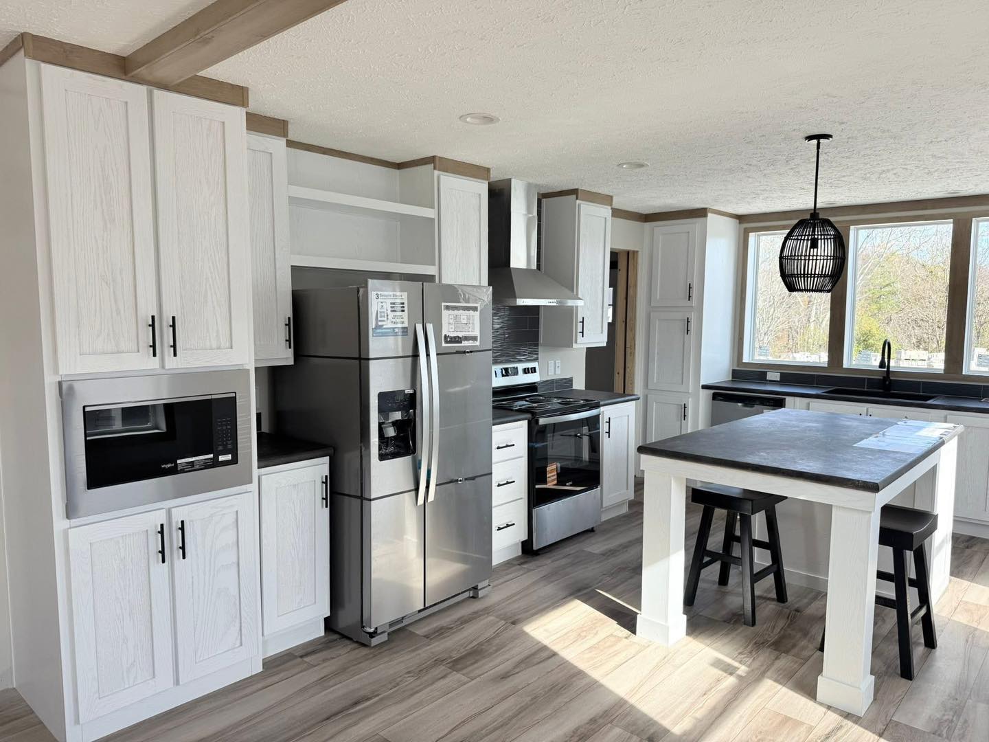 Modern kitchen with white cabinets, stainless steel appliances, and a gray island with bar stools. Large windows and pendant light create an airy, bright atmosphere.