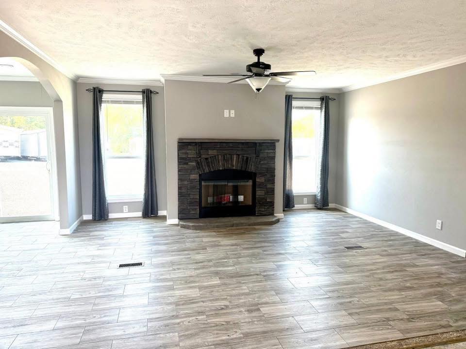 Spacious living room with gray walls, a dark stone fireplace, and light wood flooring. Two large windows with curtains and a ceiling fan enhance the airy feel.
