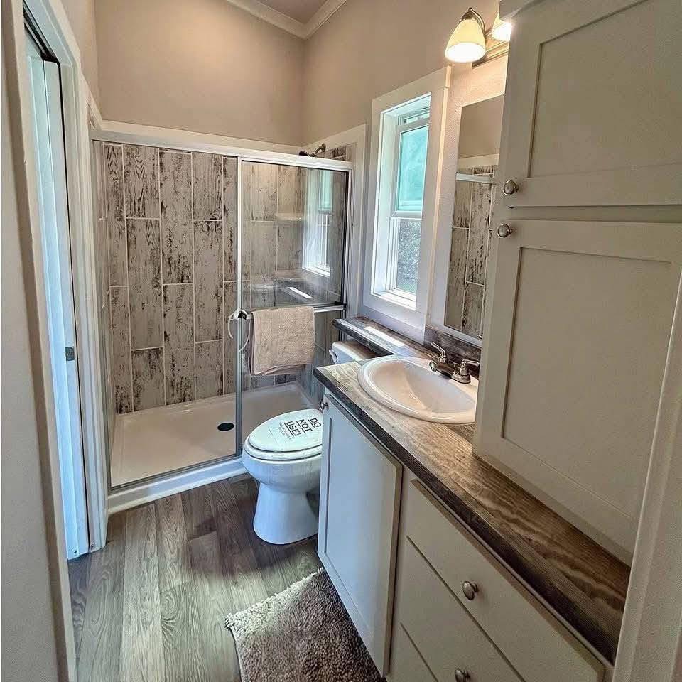 Bright bathroom with wood flooring, featuring a glass-enclosed shower with rustic tile walls, a toilet, and a sink on a wooden countertop. Warm and cozy.