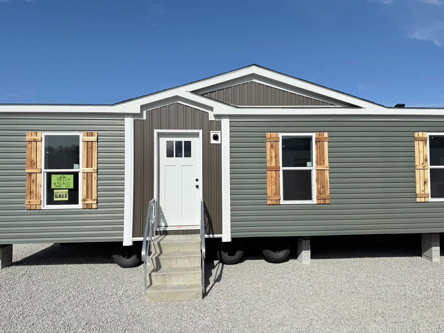 Modern mobile home with a gray exterior, wooden shutters, and a white door. A "For Sale" sign is visible in the left window. Clear blue sky.