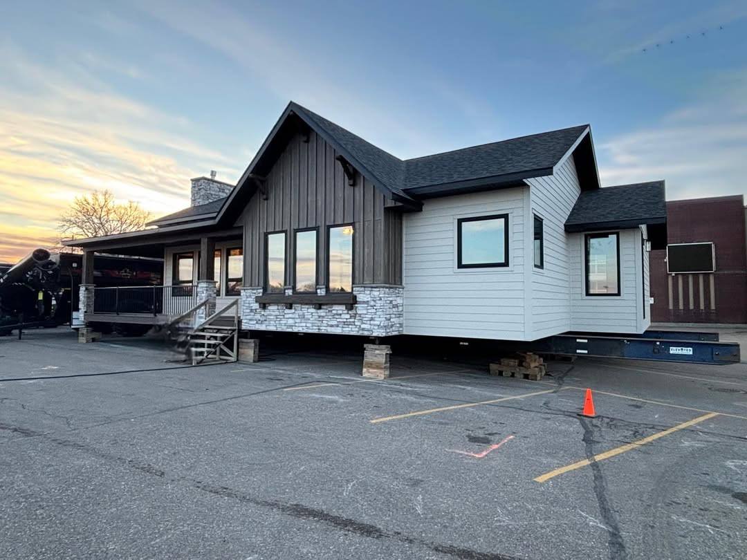 A prefabricated house with a modern design is elevated on wooden blocks in a parking lot. The sky is dusk-lit, and an orange cone is nearby.
