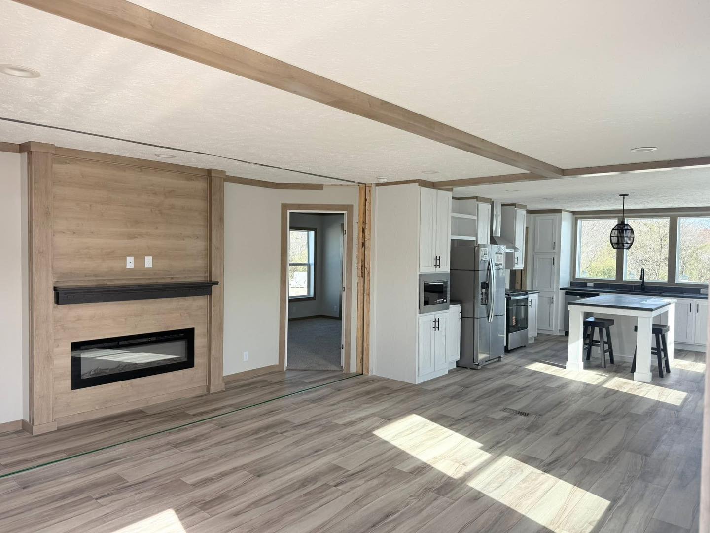 Modern open-plan living room and kitchen with light wood floors. Features include a wooden fireplace, white cabinets, sleek fixtures, and large windows.