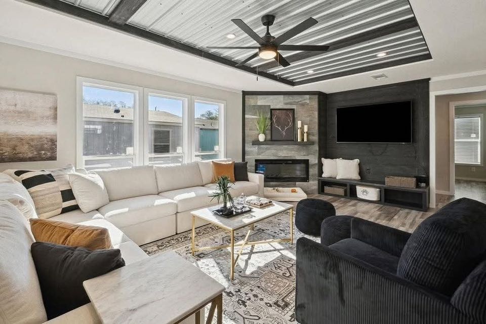 Modern living room with a white sectional sofa, black armchair, and wall-mounted TV. Features abstract art, a ceiling fan, and large windows. Cozy and stylish.
