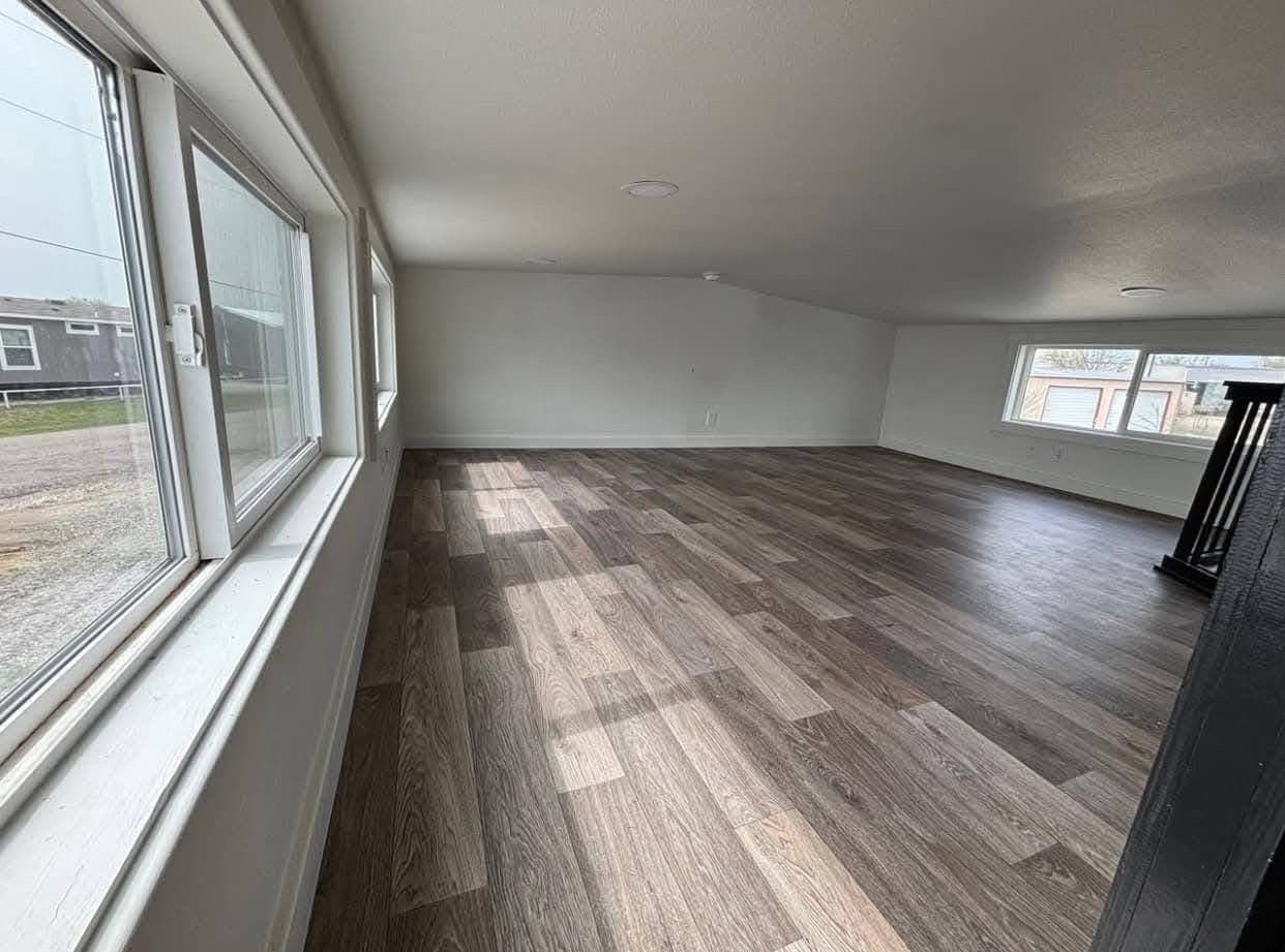A spacious, empty attic with light brown wooden flooring and angled white walls. Large windows on two sides let in ample natural light, creating a bright and airy ambiance.