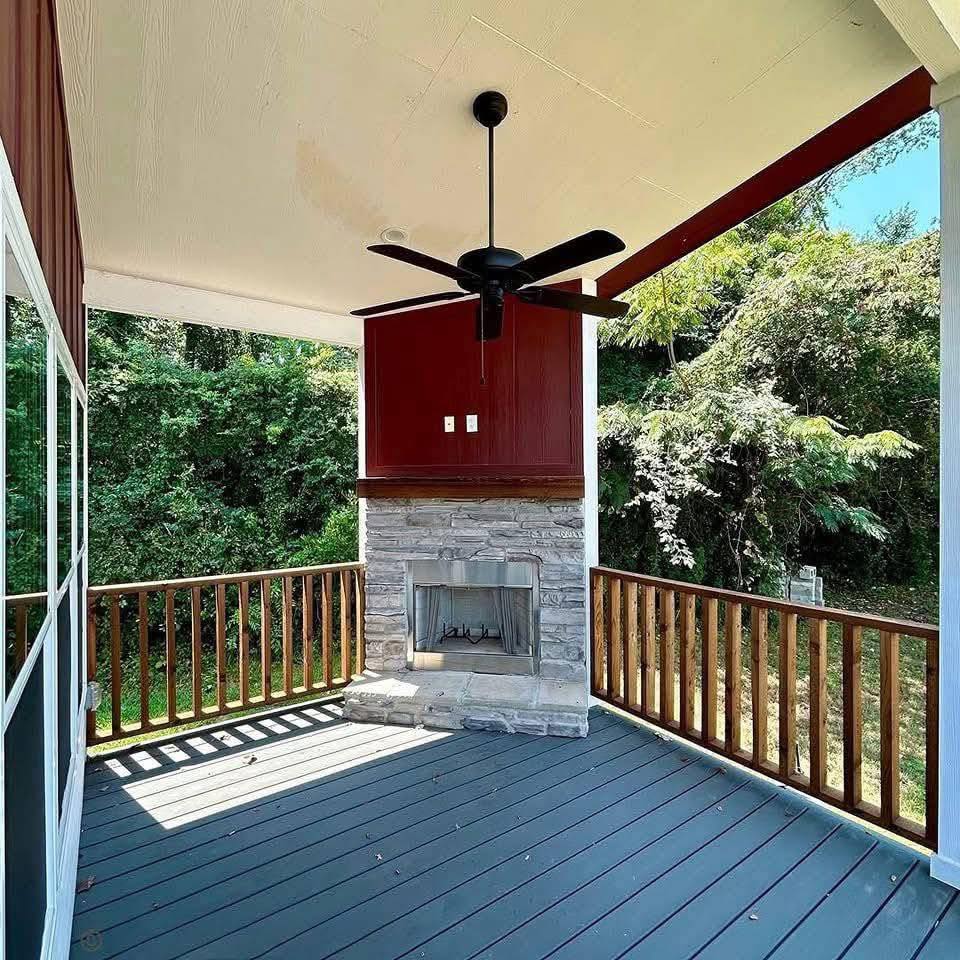 Covered porch with stone fireplace, red paneling, black ceiling fan, and wooden railing. Sunlit with lush green trees in the background. Cozy atmosphere.