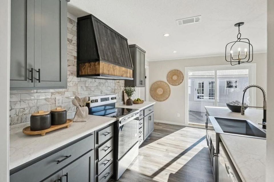 Modern kitchen with gray cabinets, marble countertops, and stainless steel appliances. Bright light from large window, warm and inviting atmosphere.