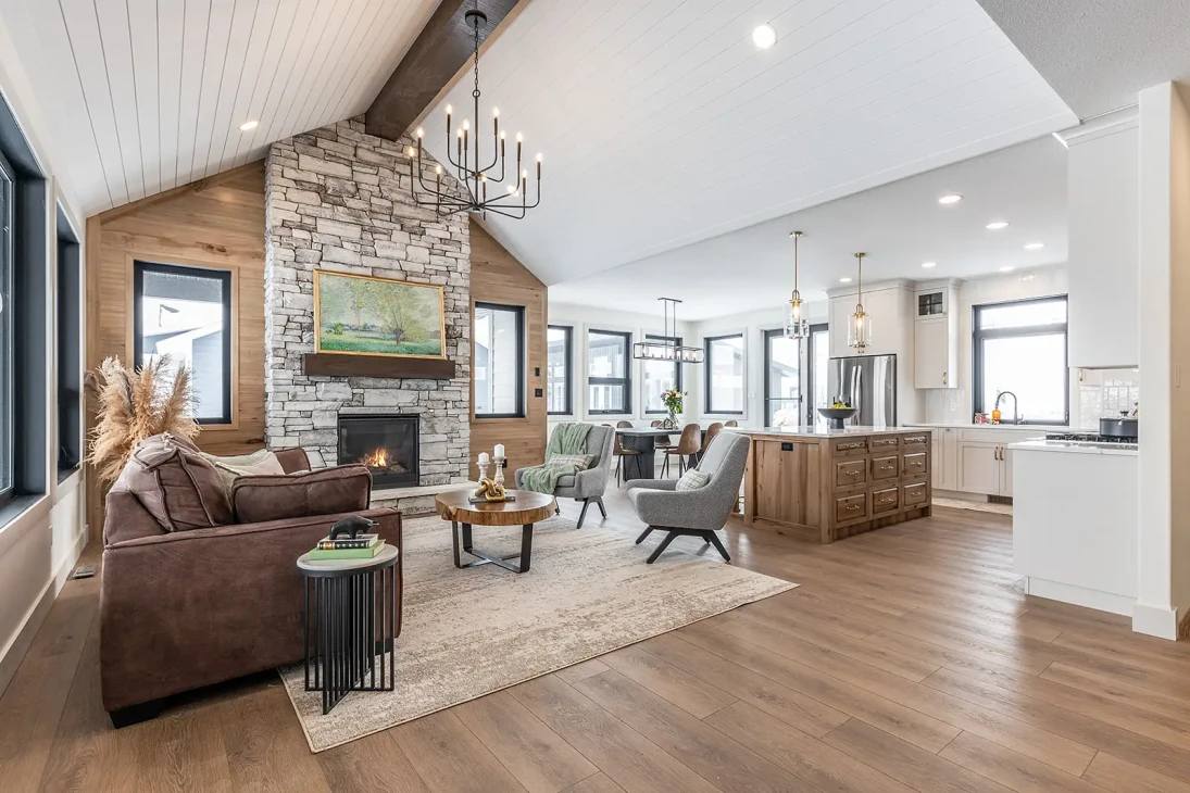 Spacious open-concept living room and kitchen with a cozy stone fireplace. Features a brown sofa, gray armchairs, wood floors, and a modern chandelier.