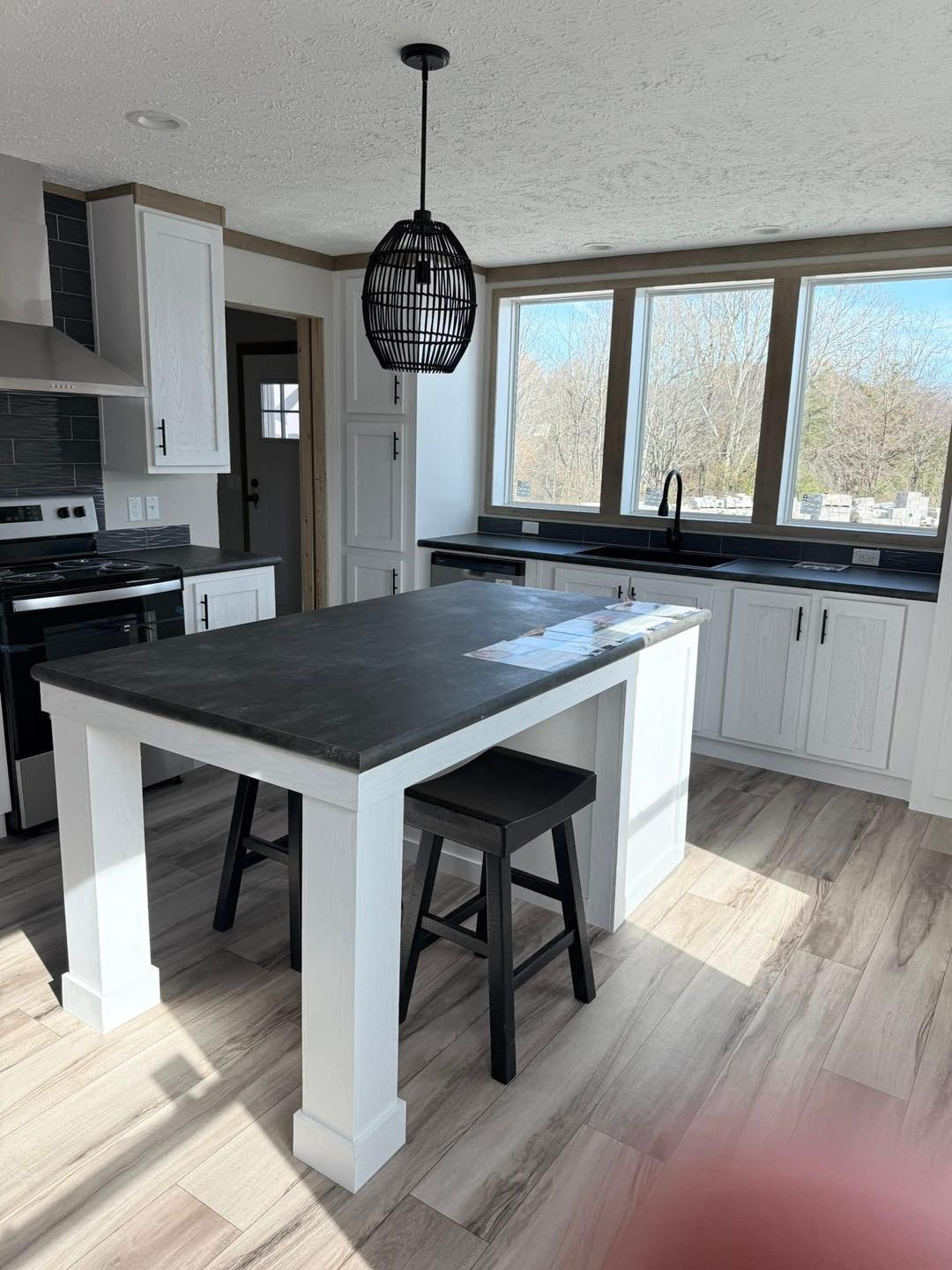 Modern kitchen with white cabinets, large windows, and natural light. Black countertop island with stools, pendant light, stove, and wood floors.