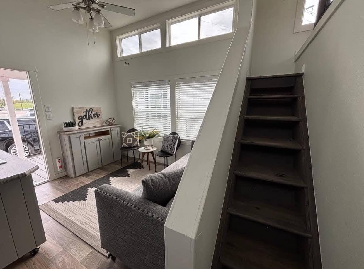 Cozy living room with gray sofa, small side table, chairs, and stairs leading upwards. Bright windows fill the space with natural light.