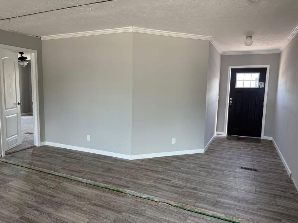 A room with light gray walls and wood-look flooring. A black front door with a window is on the right. An open doorway on the left leads to a carpeted area. The space is well-lit and minimalistic.