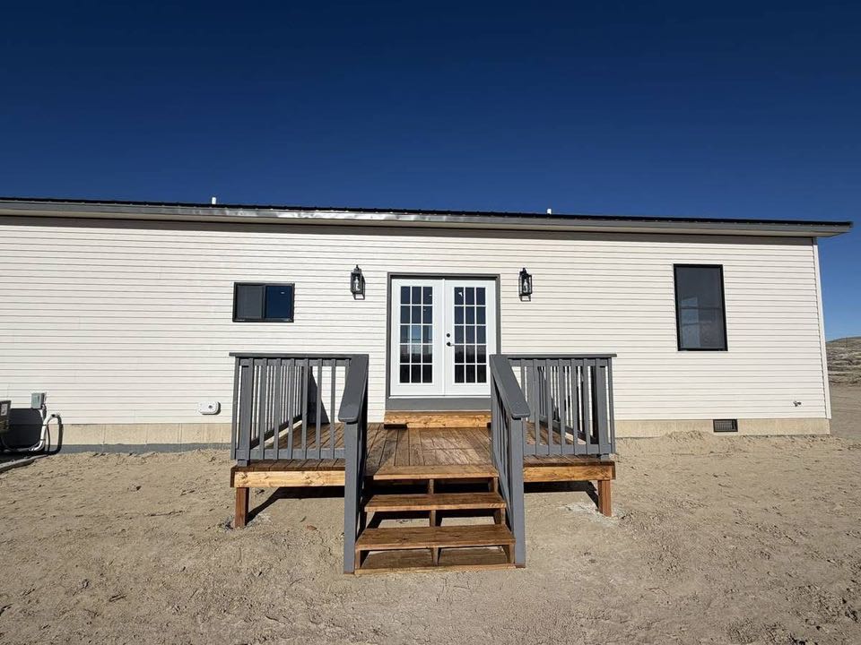 A single-story prefab home with white siding, featuring a set of wooden stairs leading to double glass doors, is set against a clear blue sky.