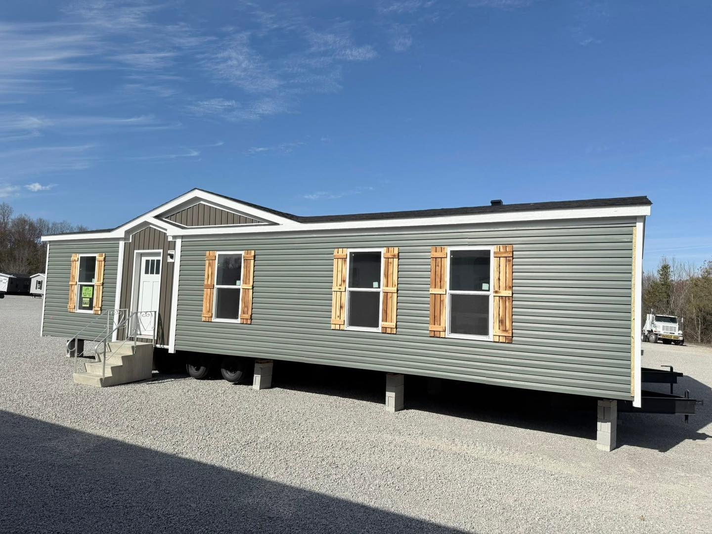 A green manufactured home with wooden shutters is set on a gravel lot. A clear blue sky is above, and trees with autumn foliage are in the background.