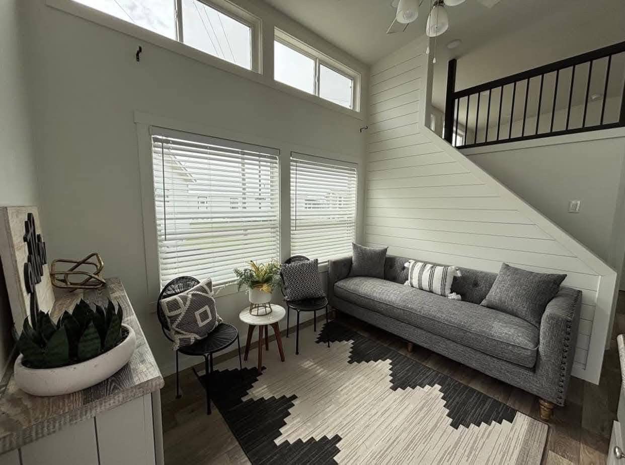 Cozy living room with a gray sofa, patterned cushions, and a geometric rug. Two wooden side tables hold plants. Large windows provide natural light.