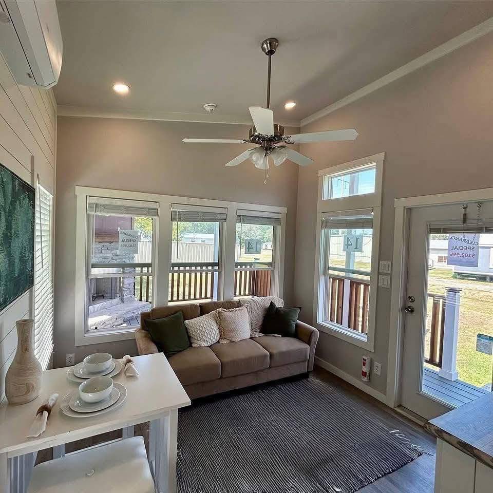 Cozy living room with a beige sofa, white and green pillows, large windows, and a ceiling fan. A small dining table with two place settings adds charm.