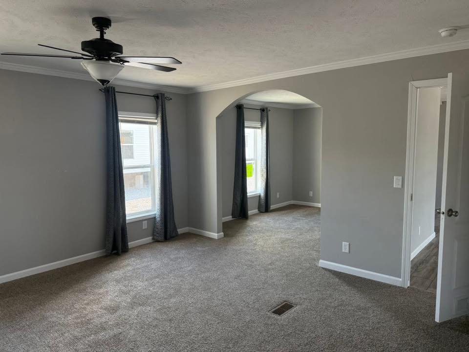 Spacious room with gray walls and carpet, a ceiling fan, and two windows with dark curtains. An arched doorway leads to another brightly lit area.