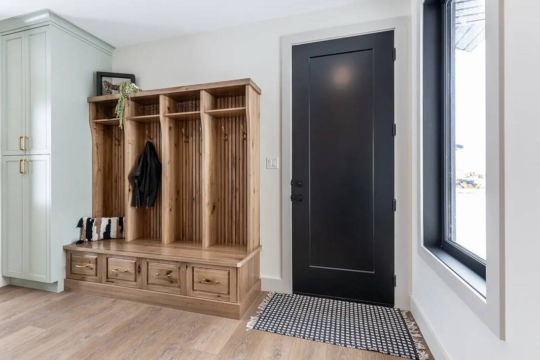 Spacious entryway with light wood flooring, featuring a wooden built-in cubby with hooks and drawers on the left, next to a black door and checkered rug.