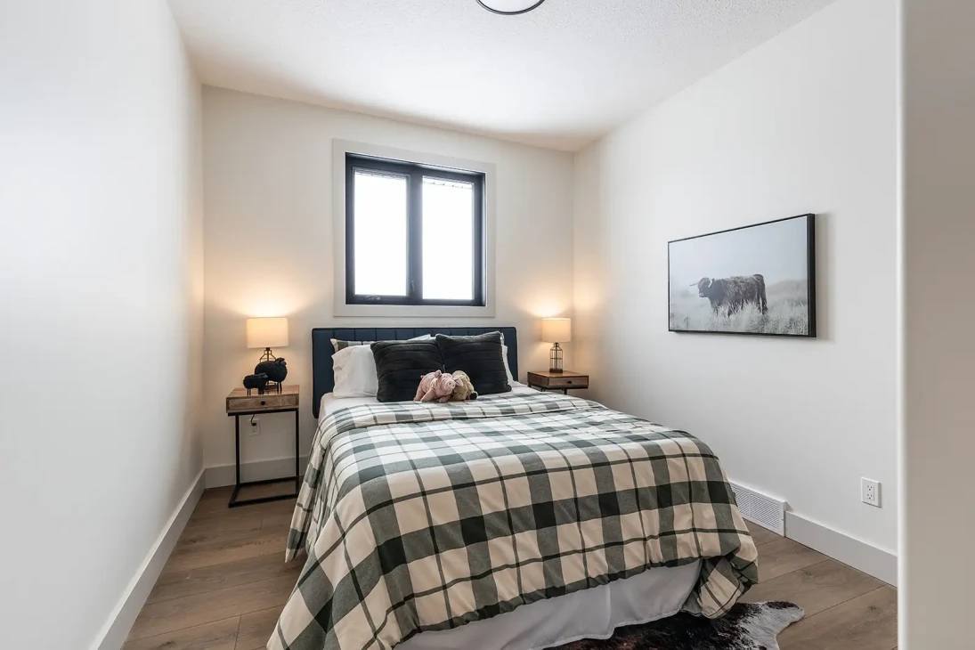 Small bedroom with a cozy feel, featuring a plaid-covered bed centered under a window. Soft lighting from bedside lamps, and a cow print on the wall.