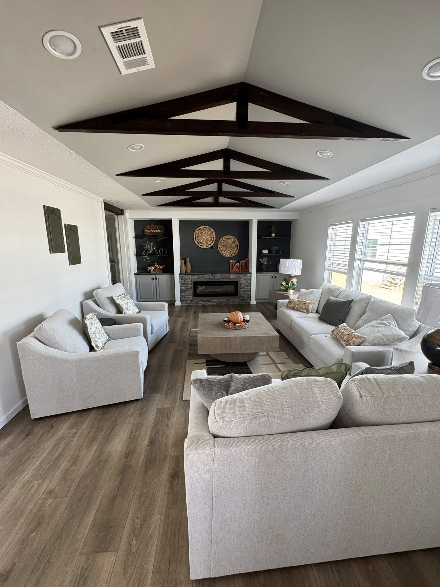 Spacious living room with light gray sofas, a central coffee table, and a modern fireplace. Exposed wood beams and large windows enhance the airy feel.