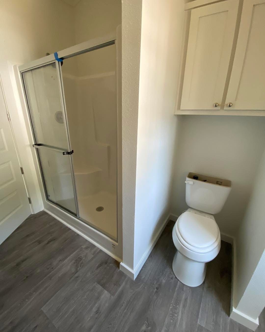 Small bathroom showcasing a glass-enclosed shower and a white toilet. The space has white walls, light wood flooring, and overhead cabinets. Clean and minimalistic design.