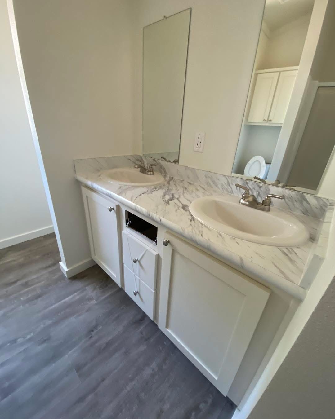 Bathroom with double sinks, marble countertop, and white cabinetry. Large mirror above, gray laminate flooring, and a glimpse of a toilet in the background.
