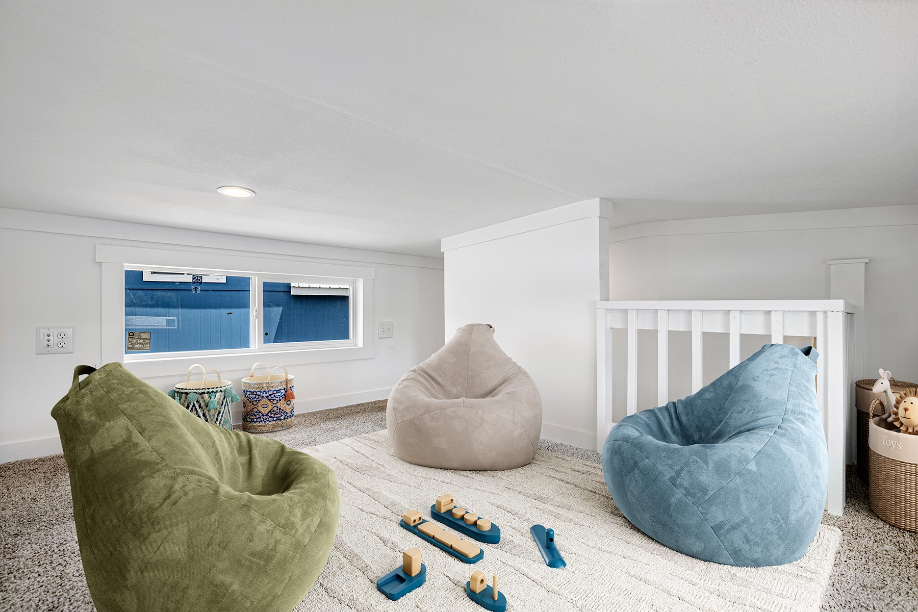 Cozy playroom with green, beige, and blue bean bags on a white carpet. Wooden toys scattered, a window with a blue view, and baskets in the corner.